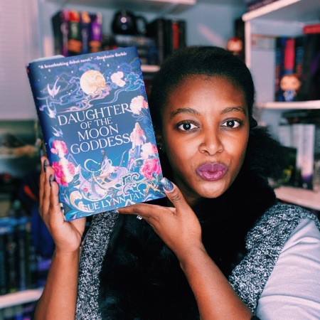 Ahtiya, a brown-skinned Black woman, is holding a hardcover copy of Daughter of the Moon Goddess.